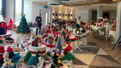 Katy Lewis/BBC A hotel room is adorned with 102 Christmas-themed knitted creations. These are essentially large hats with festive additions such as a knitted snowman or pinecone to cover the top of post boxes.