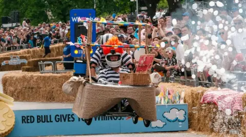 Red Bull Paul Beesley and Lee Masser wearing black and white-striped prisoner outfits while riding a large brown kart with a colourful hot air balloon on top. A large crowd watches on as they jump a ramp.