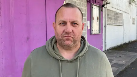 Stuart Lawrence staring at the camera wearing an olive coloured hoodie. He is standing in front of the building. The wall behind him is painted purple.