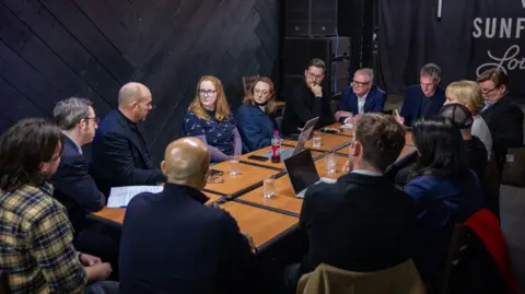 West Midlands Combined Authority Fourteen people sitting around a table, which includes glasses of water and electronic devices.