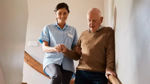 A young female careworker helping an elderly man to walk down a wooden staircase at home.   The careworker has dark brown hair tied back and is wearing a light blue nurse's tunic over dark trousers.  The man has receding white hair and is wearing a brown jumper and navy trousers. 