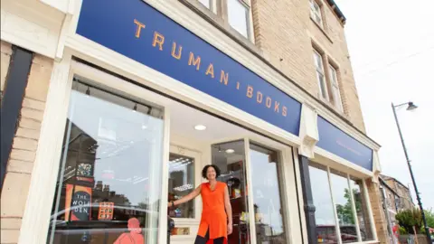 Truman Books/Supplied A woman with short dark curly hair and an orange sleeveless shirt dress and black leggings stands in front of a glass shop front.