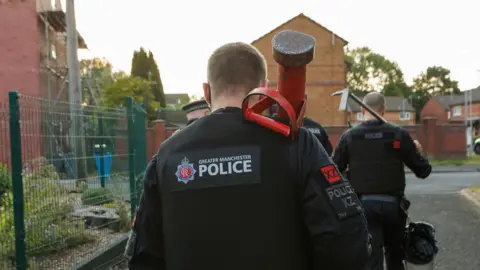 Greater Manchester Police Greater Manchester Police officers taking part in drugs raids in a residential area in Gorton. Officers are carrying tools to break down doors in the strikes.