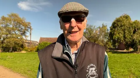 An older man with a plaid cap on his head, wearing a blue and white shirt with a brown gilet on top. There is green grass behind him and trees