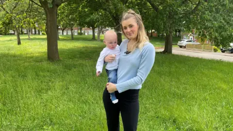 Natalie Cater holding her young son while standing on some grass next to Spinningdale in Arnold