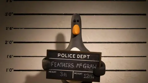 Aardman Animations Ltd / BBC Feathers McGraw, een stop-motion geanimeerde pinguïn, wordt gezien terwijl hij een bord van de politie omhoog houdt terwijl er een mugshot wordt gemaakt