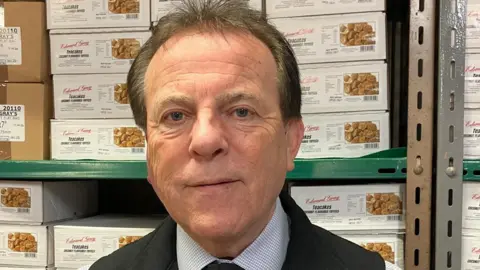 A man in a blue checked shirt is standing looking at the camera. He is not smiling and wearing a tie. Behind him are boxes of sweets stacked on shelves.