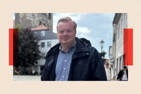 A white man with blond hair wearing a light blue shirt and a dark blue jacket smiles at the camera