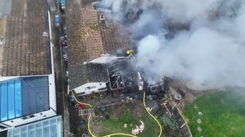 Aerial drone shot of a house fire. Billowing smoke and firefighters can be seen. 