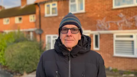 A man stood in front of a house wearing a blue stripy bobble hat and a black puffer coat. He is wearing glasses and staring at the camera. 