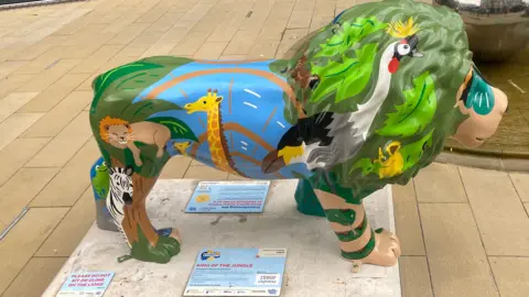 A brightly painted sculpture of a lion displayed in an outdoor public area. The lion sculpture is positioned on a white base and is decorated with colourful illustrations of animals and nature. On its body, there are painted images of a giraffe, a lion, birds, and green foliage, along with a blue section that resembles water or a landscape scene. The lion’s mane is painted with shades of green and includes a detailed bird illustration near the front. A small sign on the base reads “KING OF THE JUNGLE”, along with additional smaller text about the artwork and a note that says “PLEASE DO NOT SIT OR CLIMB ON THE LIONS.”