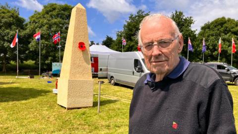 Image of Dave Addis. The temporary cenotaph and various world flags can be seen behind him. 