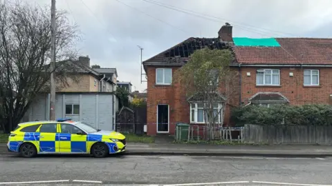 A police car outside a brick terraced house. There is a gap in the roof and rafters are exposed. 