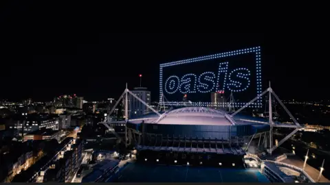 The outside of the Principality Stadium at night with high-rise buildings surrounding it. Above the stadium are purple lights in the sky which spell out Oasis, with two rectangle boxes surrounding the letters.