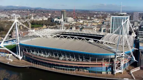 What is now the The Principality Stadium, viewed from the opposite side of the River Taff . Its four white-painted masts which hold up its retractable roof are visible. It's set against a blue sky and calm river and the city of Cardiff can be seen spreading out in the background behind it