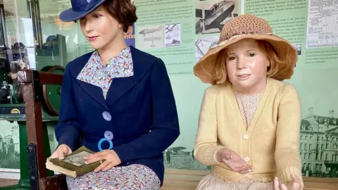 Blue Anchor Station - news and needs A child mannequin with a straw hat and cardigan and a woman mannequin wearing period clothing, sitting on a bench. There are museum displays behind them.