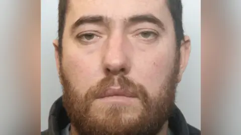 A mug shot of Viktor Ymeralilaj. He has short dark hair, a brown beard and is staring into the camera.