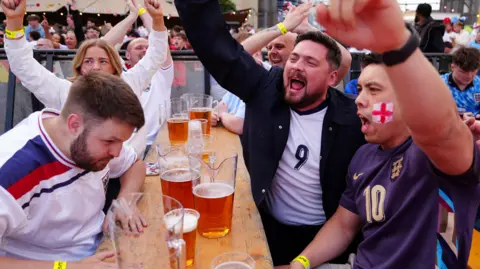 Owen Humphreys/PA A table of cheering spectators, one with an England flag painted on his cheek. The man on the left is looking down and appears to be swallowing hard. In the middle of the table are four pitchers of beer.