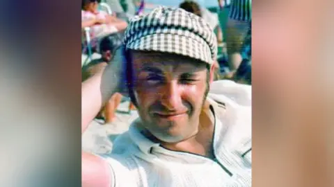 Louise Edwards A man in his forties is relaxing on a beach. He is lying on his side and is resting his head on his hand with a bent elbow. He is wearing a white-collared t-shirt and is wearing a chequered cap. It looks like a sunny day. 