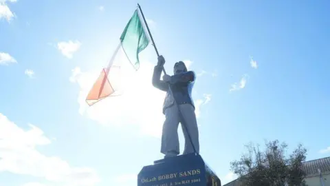 Members of the public attend the unveiling of the Bobby Sands statue. The statue is in the centre of the photo with crowds of people standing around it. It is a sunny day. 