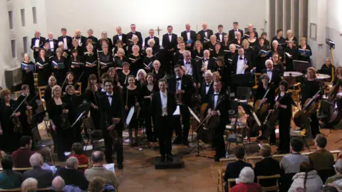 Welwyn Garden City Orchestra and Chorus A large choir and orchestra stand at the front of a modern church or concert hall during a live performance. Dozens of singers dressed in black are arranged in tiered rows behind musicians holding string instruments. A conductor stands at the centre facing the audience. Rows of seated audience members fill the foreground, while high white walls and bright stage lighting frame the performance space