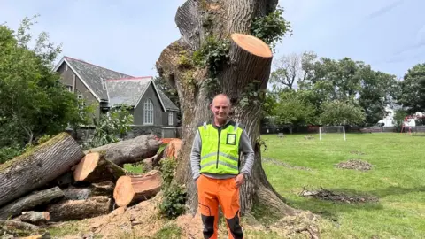 Steve Curphy stands with his hands in his pockets in front of a large trunk of an elm tree. He is wearing a bright hi-vis top with orange trousers.