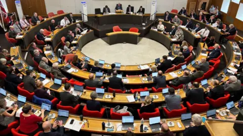 Lee Trewhela/LDRS The Cornwall Council chamber with desks and seats arranged in a circle around the room. The photographer has taken the shot from the back of the room and we can see  microphones, laptops and paperwork on councillors' desks.