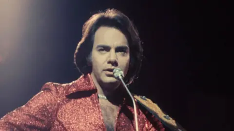 Neil Diamond sings into a microphone. He has long brown hair and wears a red suit.