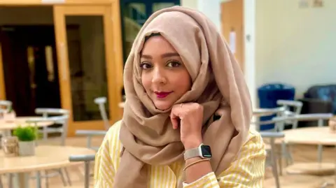 Tasneem Abdur-Rashid A woman wearing a brown hijab and yellow and white striped top smiling, she has her chin resting on her hand and she has a watch on her wrist. 