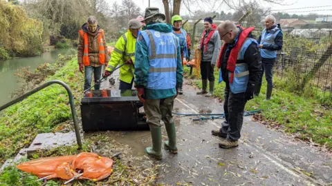 Grantham RiverCare A group of litter pickers wearing blue, yellow and orange high vis vests, with some standing on top of a wall and others sitting below it. They are holding litter picks and bags and there is also a golden retriever.