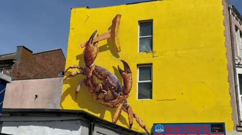 A wall with a yellow covering, and a painting of a crab which is holding a wooden fork and a chip