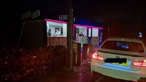 A white BMW parked outside Wing Shed in London Apprentice, near St Austell, at night. Four people are inside the roadside chicken restaurant and takeaway.