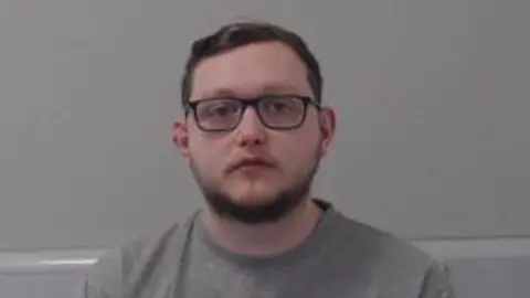 Shows a man in his 20s with a grey round-neck jumper and wearing glasses.