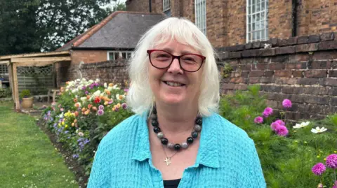 A woman with white hair and a pale blue cardigan in a garden with pink flowers