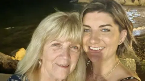 Collect/PA Real Life A recent photo of Smith and her mum. On the left, Margaret has blonde hair and a full fringe, with gold hoop earrings. On the right, Smith has short blonde-brown hair and is smiling, taking the selfie of them both. It is nighttime and in the background there looks to be a beach.