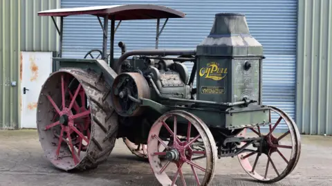Cheffins A 1917 Rumely Oil Pull tractor parked in front of a warehouse-type building.