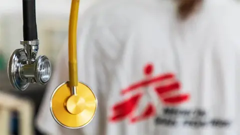 Two stethoscopes hang in the foreground in front of a medic wearing scrubs with a blurred MSF logo.