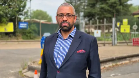 Stuart Woodward/BBC A man standing at Wethersfield in a grey/black blazer and a blue and white striped shirt. He is wearing black framed glasses and looking directly at the camera but he is not smiling. There are metal gates behind him.