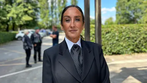 BBC News A woman dressed smartly in a black pin-striped jacket, matching cravat and white shirt. Her black hair is tied tightly in a pony tail.