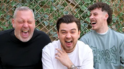 Paul, Harrison, and Travis, all laughing, in their back garden, in front of a trellis wall. Paul has short grey hair, with a beard, Harrison, dark hair and stubble and Travis, stubble, and a mullet style hair cut with the sides shaved. Paul has his eyes closed and is wearing a black top; Harrison a white T-shirt and Travis a blue top. 