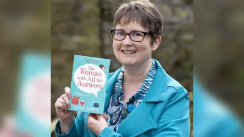 East Yorkshire Riding Council Linda Green smiling at the camera holding her book. She has short brown hair and is wearing glasses and a teal jacket over a patterned blue shirt. She is holding a book titled The Woman with All the Answers by Linda Green. The background is blurred and features an outdoor setting with a stone wall.