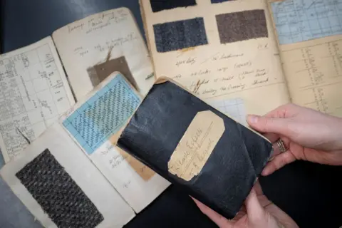 Kirsty Anderson/Lovat Mill Four books with bits of fabric pasted inside. The pages of the books are yellowing and have handwriting in them, though it is not clear what it says. 