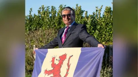 Dal Roy Dal Roy is holding a Fenland flag in both hands while stood outside. He is wearing a suit with a pink tie and is wearing sunglasses. 