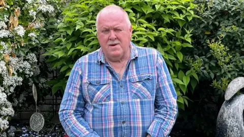 Julia Gregory / BBC A man wearing a blue, pink and white shirt. He is sitting in a garden with his hands in his lap.