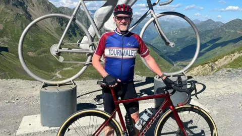 John Ireson John Ireson with his cycling gear on. He is wearing a lycra, helmet and glasses. There is a picturesque behind him with mountains and valleys. 