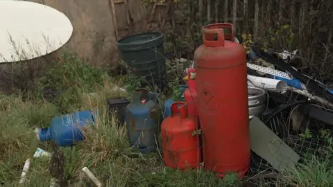 Fiona Irving / BBC A pile of blue and red gas canisters. They are in a field with overgrown rubbish, surrounded by other rubbish.