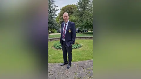 LDRS A man wearing a suit with a navy and red tie. He has his hands in his pocket and it standing in the gardens. 