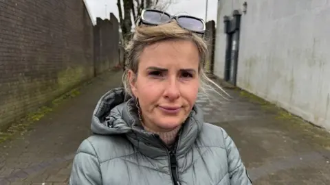 Stacey White, a woman with blonde hair with sunglasses on her head and wearing grey raincoat standing in the streets of Rhymney looking straight ahead