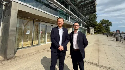 Councillors Chris Eynon, on the left, and Andrew Ramsey, both wearing black suit jackets and open-buttoned shirts, stand in front of the now closed National Glass Centre in Sunderland. The building behind them is boarded up and there are large wooden panels behind the glass-fronted entrance. 