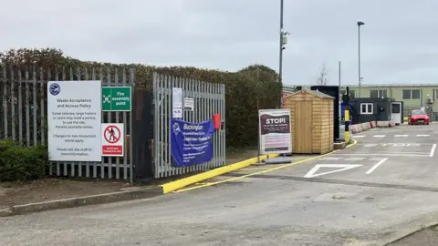 Buckinghamshire Council An entrance to a recycling site, with a wooden shed, iron gates that are open, a cabin in the distance, several signs on the gates and a road with markings on it.